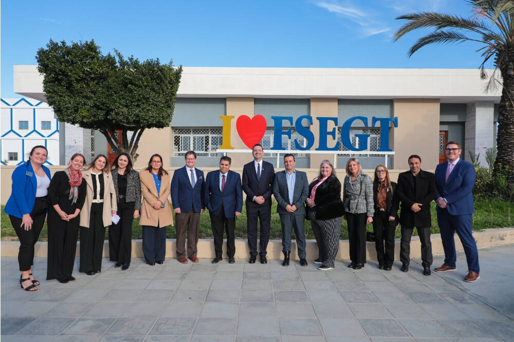 GVSU officials dressed in business and professional attire stands in a line outside in front of a building with a large sign that reads "I ♥ FSEGT." The scene features clear blue skies, a palm tree, and a trimmed tree.
